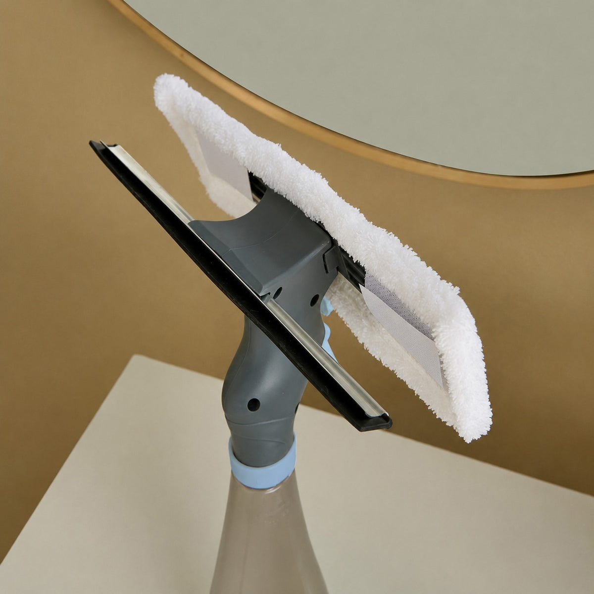 A close-up of a window cleaning squeegee attachment with a white microfiber cloth and a gray plastic handle, mounted on a spray bottle. The background shows a warm beige wall with a partial view of a gold-framed mirror.