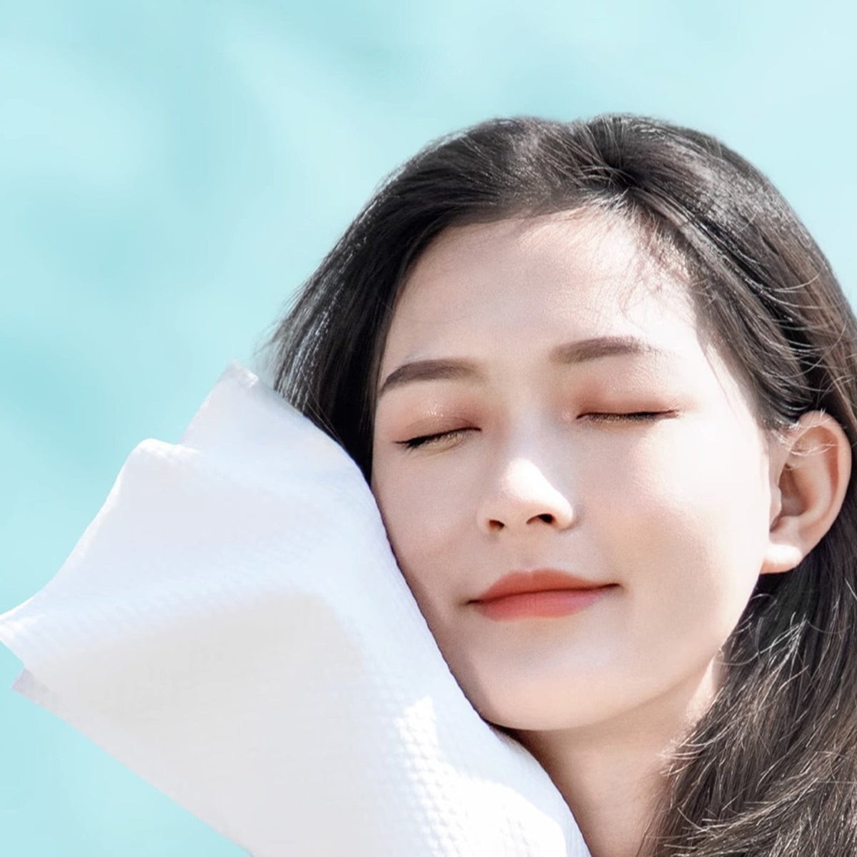 A close-up lifestyle shot of a woman with closed eyes resting her head on a compression bath towel, set against a soft, light blue background.