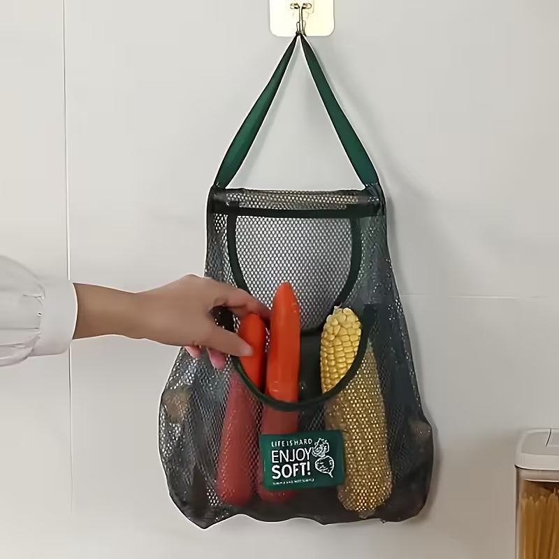A mesh hanging storage bag attached to a wall hook, with a hand placing vegetables inside. The bag has a large main compartment and a front pocket, used for storing items like carrots and corn while keeping them visible and ventilated.
