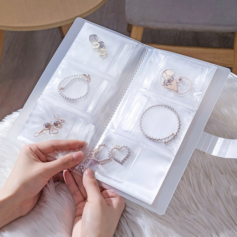 A person is holding  transparent jewelry storage book with multiple clear pocket sleeves displaying neatly organized earrings, bracelets, and rings.