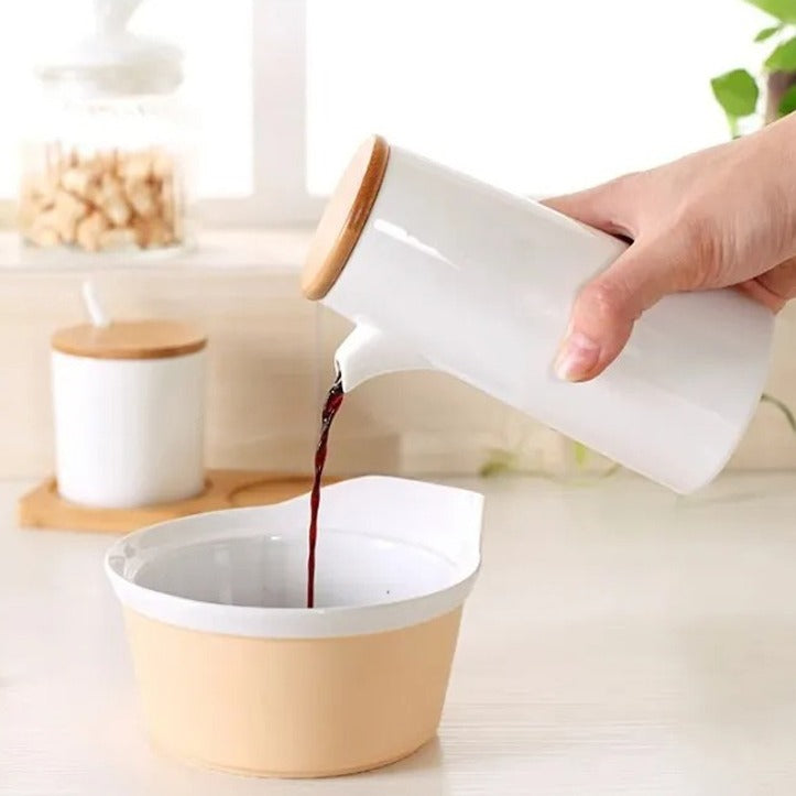 A person pouring liquid from a ceramic bottle into a bowl in a kitchen, with a matching seasoning container set placed on a wooden stand nearby.