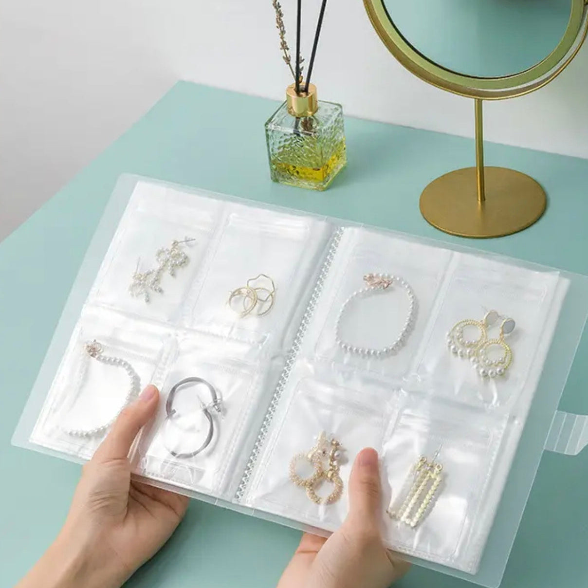 A Person is holding a transparent jewelry storage book with multiple clear pocket sleeves displaying neatly organized earrings, bracelets, and rings.